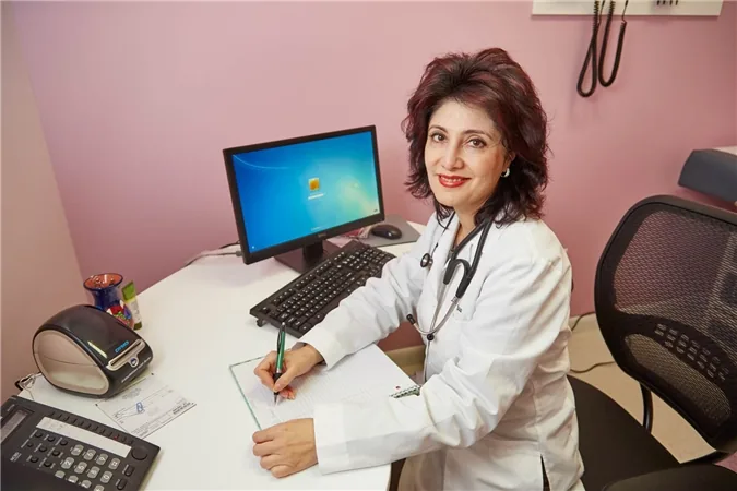 La Dra. Irina Ilyayeva con bata blanca y estetoscopio, escribiendo en su escritorio del consultorio